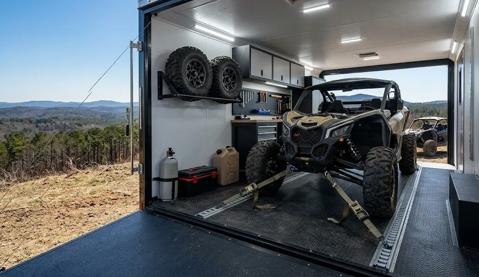 An interior view of a modern enclosed trailer containing a secured UTV, spare tires on a wall rack, and a organized workbench with tools.