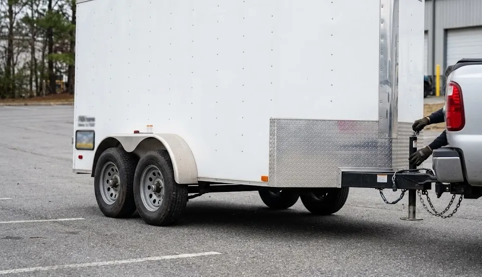 A white tandem-axle enclosed cargo trailer hitched to a silver pickup truck in a parking lot.